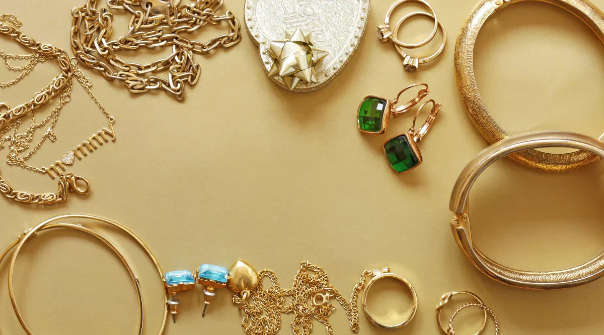 A flat lay of various gold-filled jewelry pieces including necklaces, rings, bangles, and earrings with green and blue gemstones, arranged on a golden background.