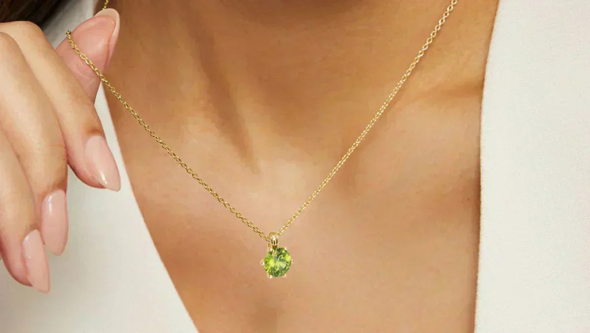 Close-up of a woman- wearing- a gold -chain necklace with a round-cut green peridot pendant, gently holding the chain with manicured fingers.