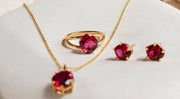 Gold jewelry set featuring a matching ruby pendant necklace, ruby ring, and ruby stud earrings, displayed on a soft beige surface.
