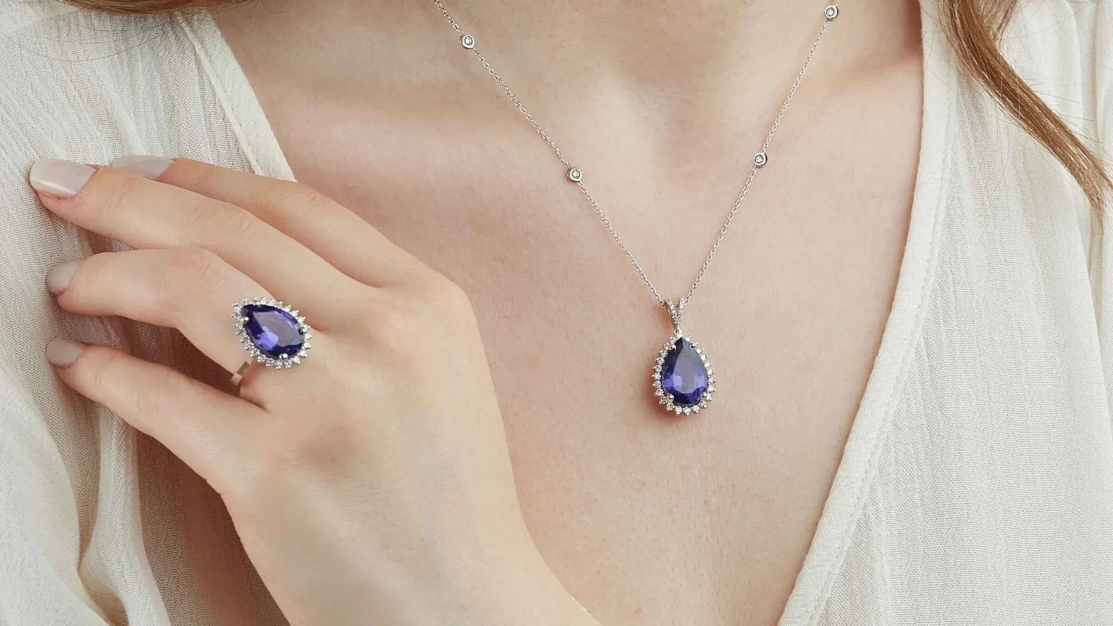 Woman wearing a matching pear-shaped blue sapphire ring and necklace set, both surrounded by small white gemstones, styled with a white outfit.