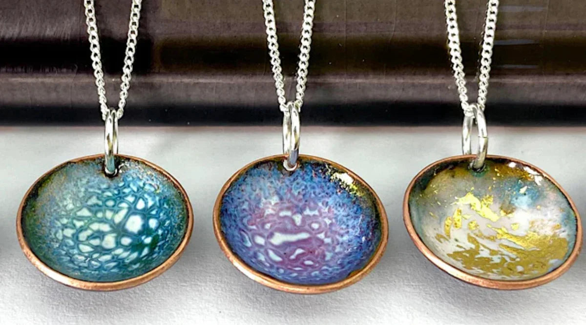 Three handcrafted enamel jewelry pendants with colorful glass-like finishes in blue, purple, and gold tones, each set in copper and hanging from silver chains. 