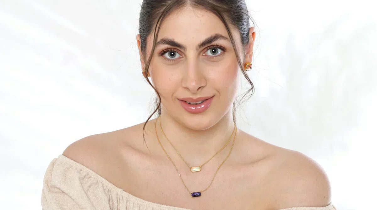 Woman wearing a beige off-shoulder top paired with gold-toned gemstone earrings and layered rectangular stone pendant necklaces, posing against a soft white background.