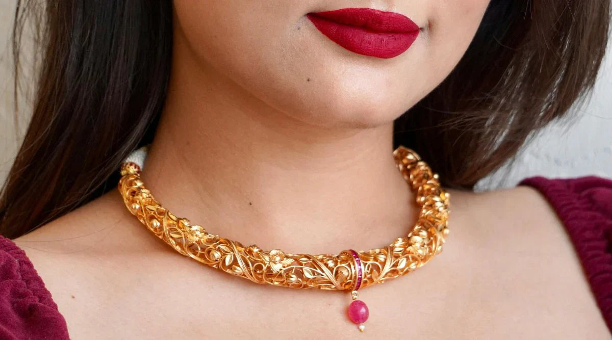 Close-up of a woman wearing a traditional, intricately designed gold necklace with a pink gemstone pendant, paired with bold red lipstick and a maroon outfit.
