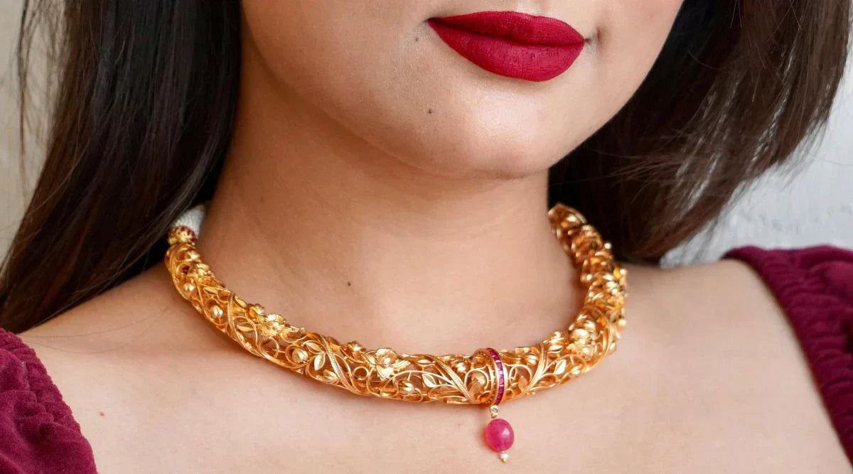 Close-up of a woman wearing a traditional, intricately designed gold necklace with a pink gemstone pendant, paired with bold red lipstick and a maroon outfit.