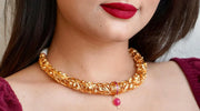 Close-up of a woman wearing a traditional, intricately designed gold necklace with a pink gemstone pendant, paired with bold red lipstick and a maroon outfit.
