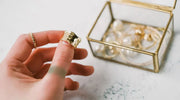 Close-up of a hand holding a shiny gold-plated ring near a glass jewelry box filled with gold and pearl accessories on a light surface.