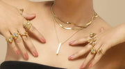 Close-up of a woman's hands and neck adorned with multiple gold-plated rings and layered gold-plated necklaces, featuring intricate designs including hearts, butterflies, and geometric shapes.