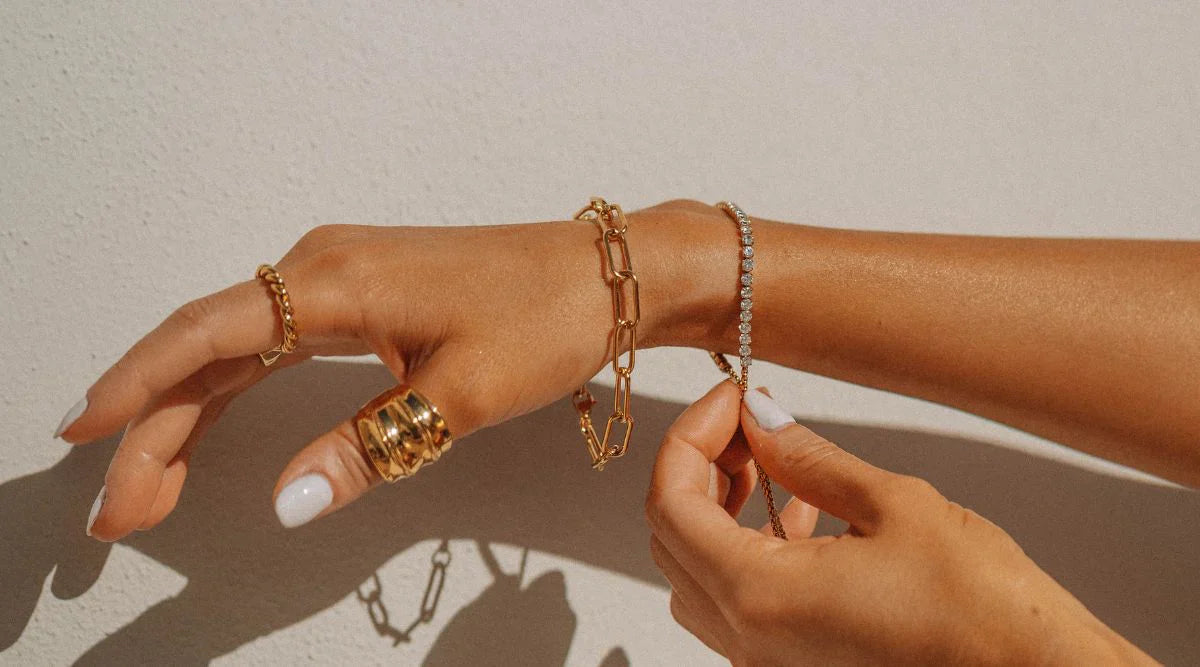 A close-up of a woman’s hands wearing multiple gold-plated rings and bracelets, adjusting a delicate chain bracelet under natural sunlight against a light background.