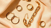 A collection of gold-plated jewelry pieces including hoop earrings, rings, and a chain bracelet displayed on a soft beige fabric background.