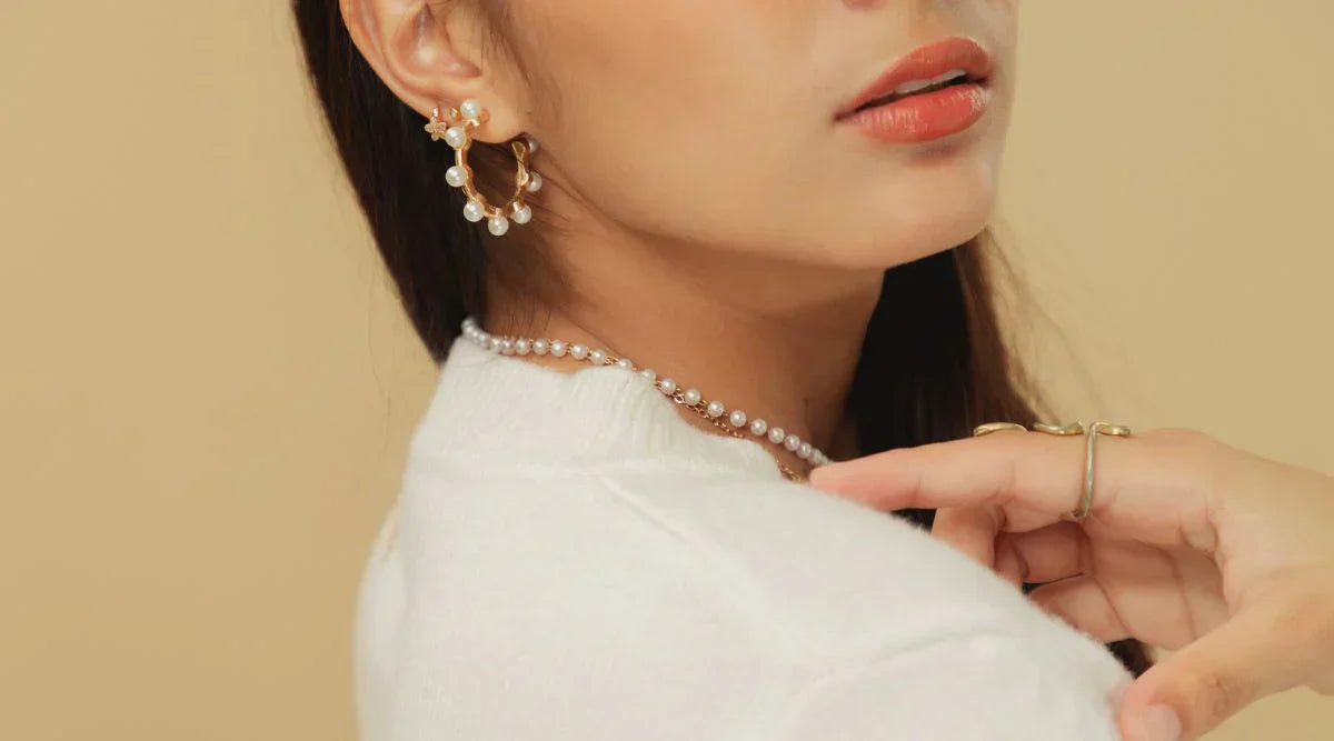 Close-up of a woman wearing elegant pearl hoop earrings, a pearl necklace, and gold rings, styled with a white outfit against a beige background.