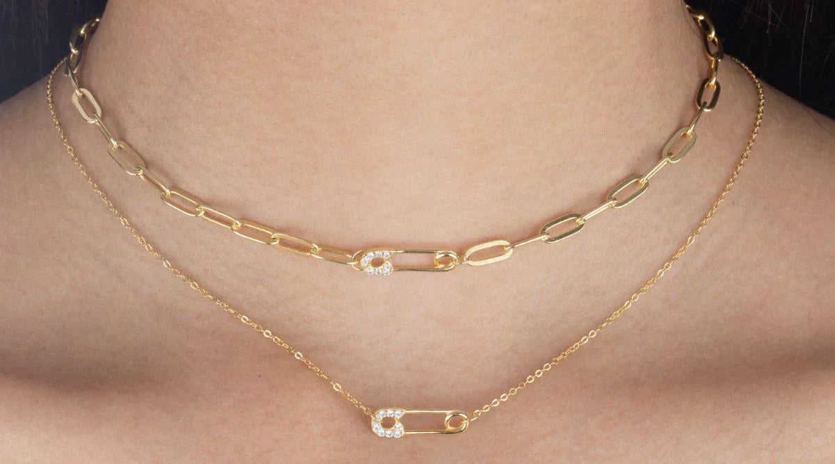 Close-up of a woman's neck wearing two gold safety pin necklaces one with a chunky chain and the other with a delicate chain both featuring diamond studded safety pin pendants.