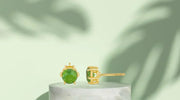 Gold-plated stud earrings featuring round green peridot gemstones, displayed on a marble pedestal with a soft green background and leaf shadows.
