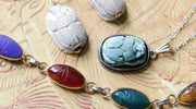 Assorted scarab-shaped jewelry pieces including carved stone amulets, a pendant on a chain, and a colorful linked bracelet featuring scarab designs in blue, red, purple, and green, displayed on an ancient Egyptian-style background.