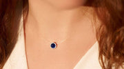 Close-up of a woman wearing a delicate necklace with a round blue sapphire pendant surrounded by small white stones, resting on her neck.