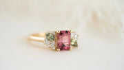 Elegant gold ring featuring a rectangular pink spinel gemstone in the center, flanked by green gemstones and small white diamonds arranged in a floral cluster design, displayed on a soft cream background.