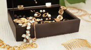 Open black jewelry box displaying assorted gold and pearl earrings, bracelets, and rings, with additional gold accessories arranged on a light wooden surface.