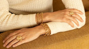 Close-up of a person wearing gold-plated bracelets and floral gold rings on both hands, resting on a textured brown surface while wearing a cream-colored sweater.